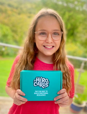 Young girl holding a blue 'Hero Cards' box outdoors with greenery in the background