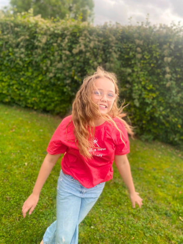 Girl wearing a red shirt and blue jeans standing in a grassy area with green bushes in the background