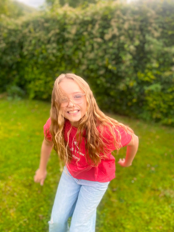 Young girl in a red shirt and blue jeans standing in a grassy area with greenery.